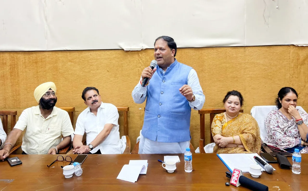 Mayor Vikas Sharma speaking into a microphone at the Nagar Nigam hall, officials and traders seated beside him