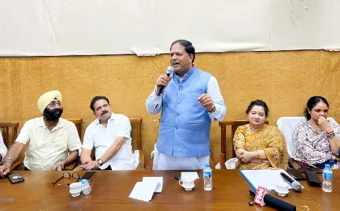 Mayor Vikas Sharma speaking into a microphone at the Nagar Nigam hall, officials and traders seated beside him
