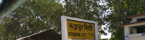 रुद्रपुर सिटी रेलवे स्टेशन का पीला नामपट्ट, जिस पर हिंदी, अंग्रेज़ी और उर्दू में स्टेशन का नाम लिखा है।
